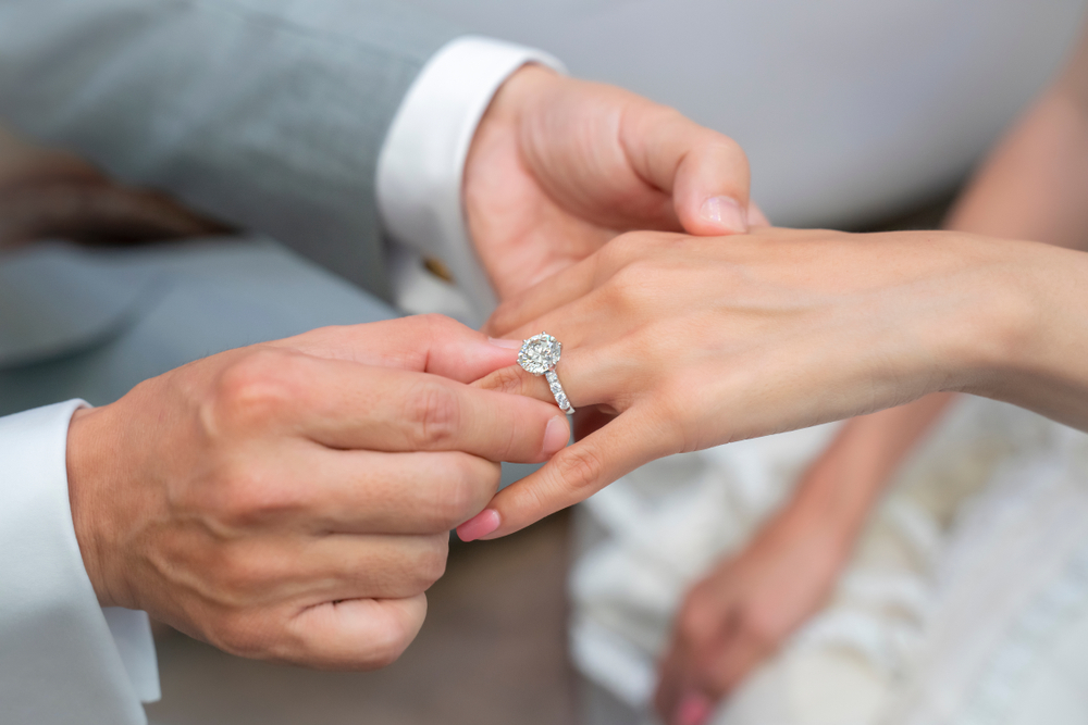 white male hands putting on a large diamond ring onto a woman's hand with pink nails